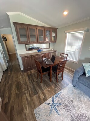 A dining area with a wooden table and chairs, a kitchen with wooden cabinets, and a window with blinds. - Snowbird Retreat in Beautiful Bradenton Florida! (Bradenton)