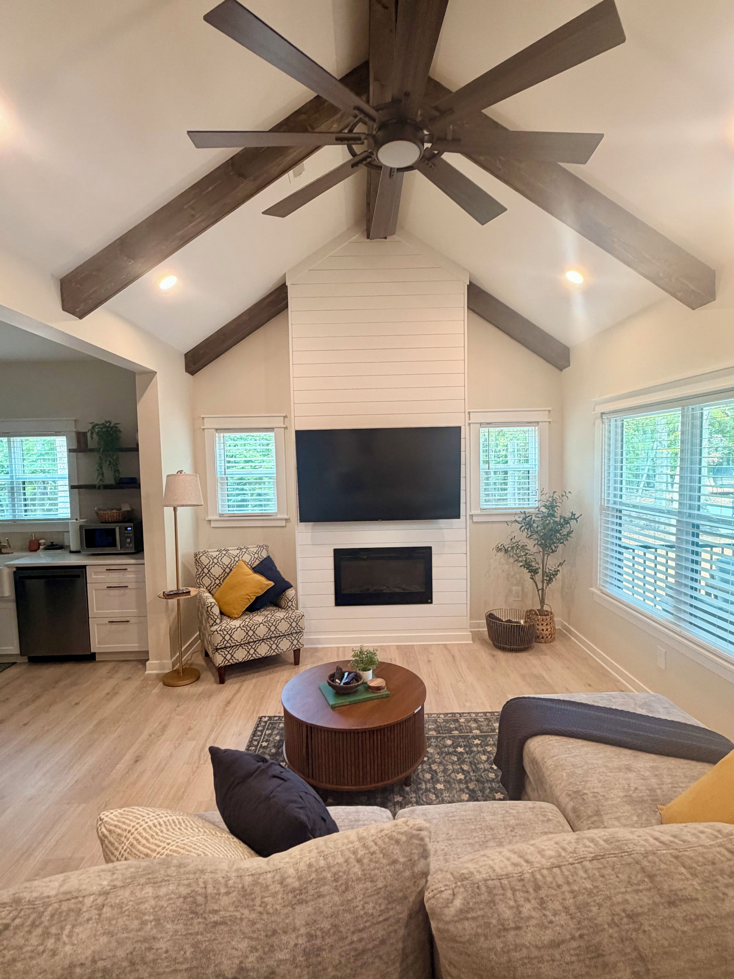 A spacious living area with a high ceiling, a fireplace, a flat-screen TV, and a kitchen in the background.
