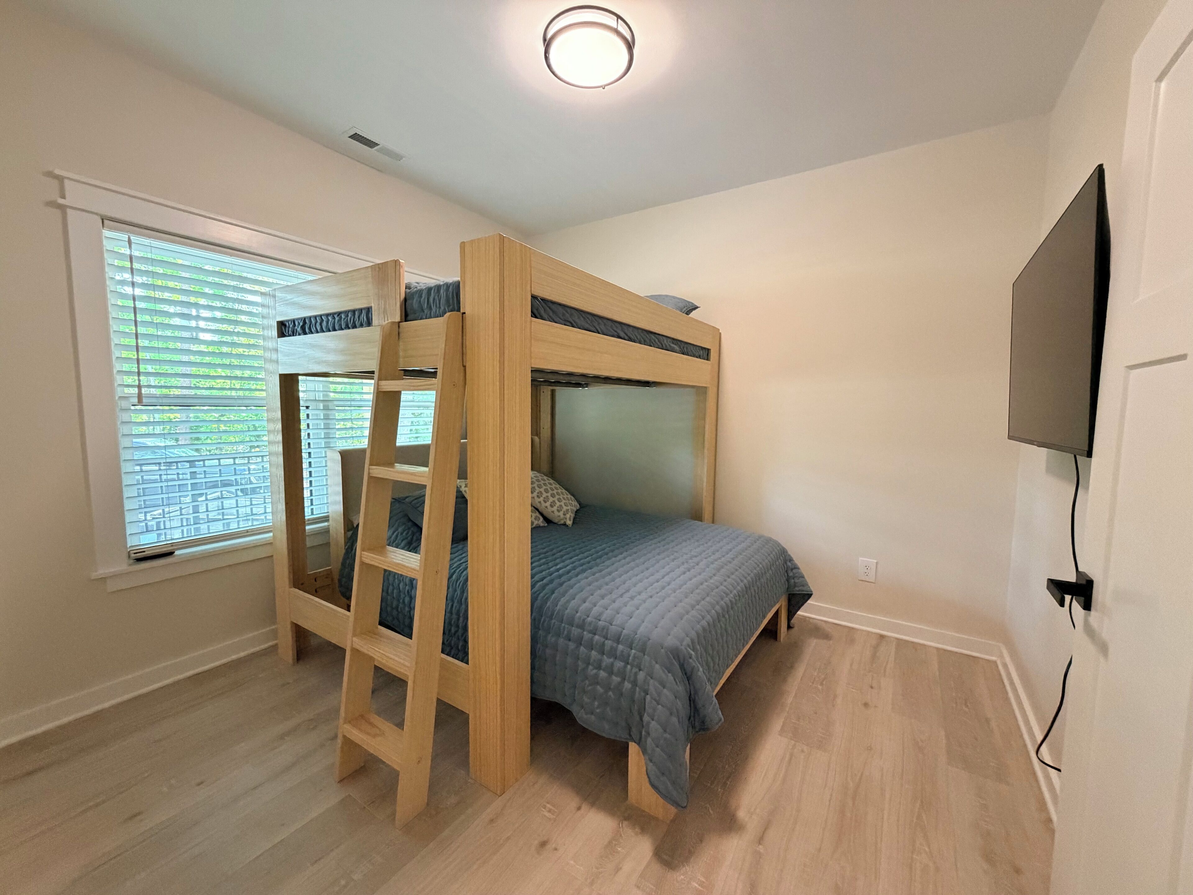 Een stapelbed met een ladder, een raam met zonwering, en een aan de muur bevestigde televisie.