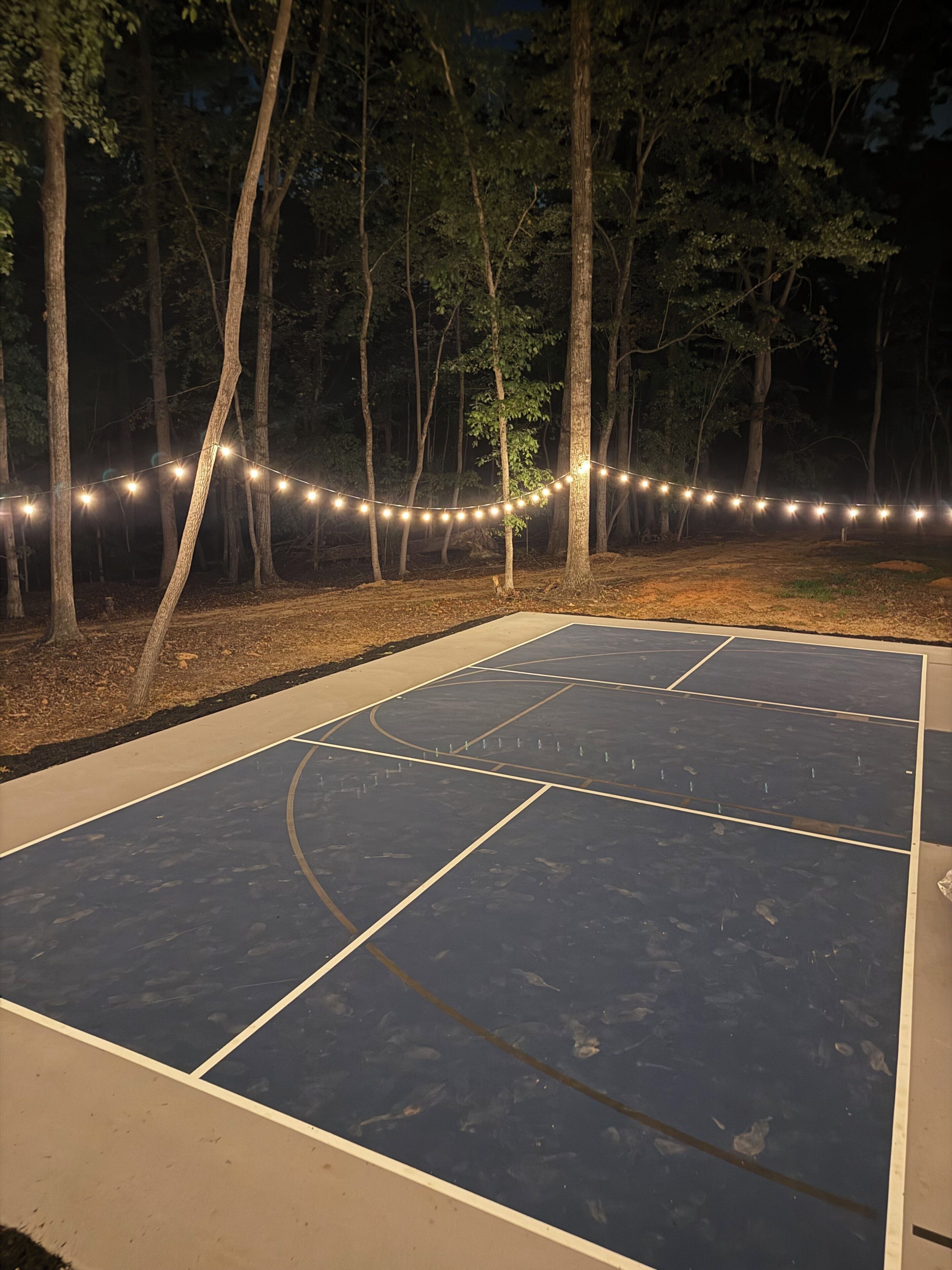 An outdoor basketball court with a wooden fence and string lights.