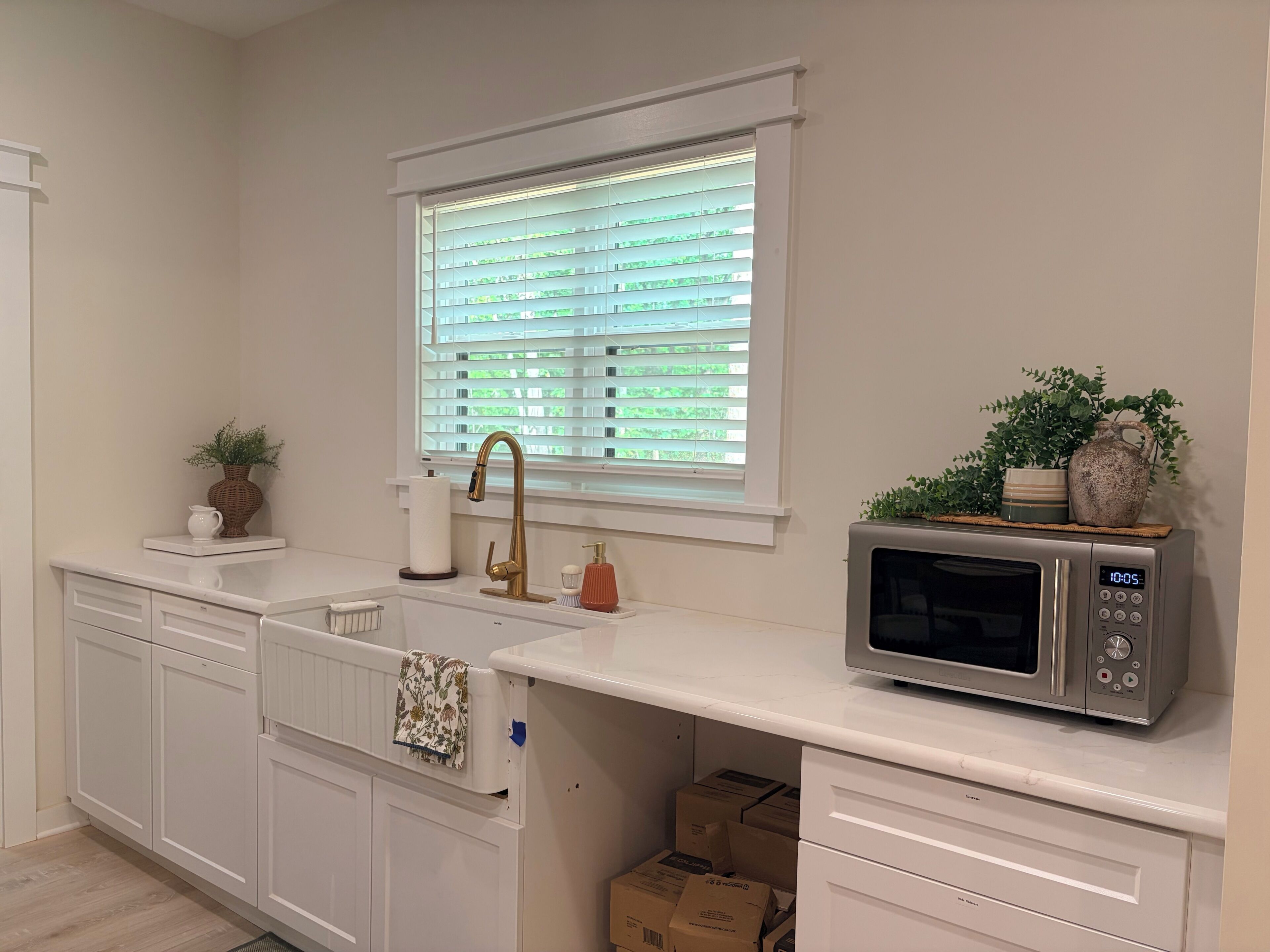 A kitchen with white cabinetry, a stainless steel microwave, a window with blinds, and a sink with a gold faucet.