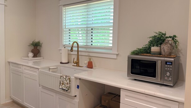 A kitchen with white cabinetry, a stainless steel microwave, a window with blinds, and a sink with a gold faucet.