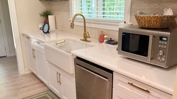 Een moderne keuken met een RVS vaatwasser, een magnetron en een gootsteen met een gouden kraan. Daarboven bevinden zich houten planken en een raam met uitzicht op groen.