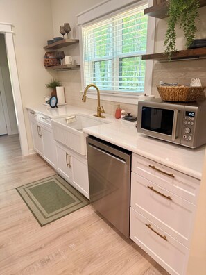 A modern kitchen with a stainless steel dishwasher, a microwave, and a sink with a gold faucet. There are wooden shelves above the sink and a window with a view of greenery outside.