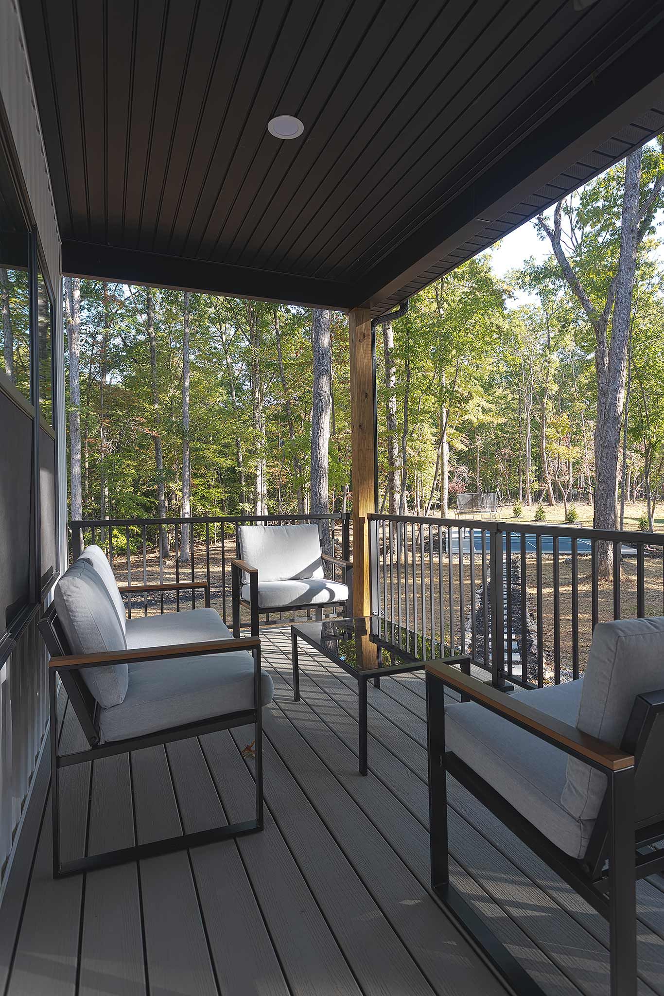 Un espace extérieur aménagé, abrité et doté de deux chaises et d’une table, entouré d’une terrasse en bois et d’une rambarde en métal, offrant une vue sur une zone boisée.