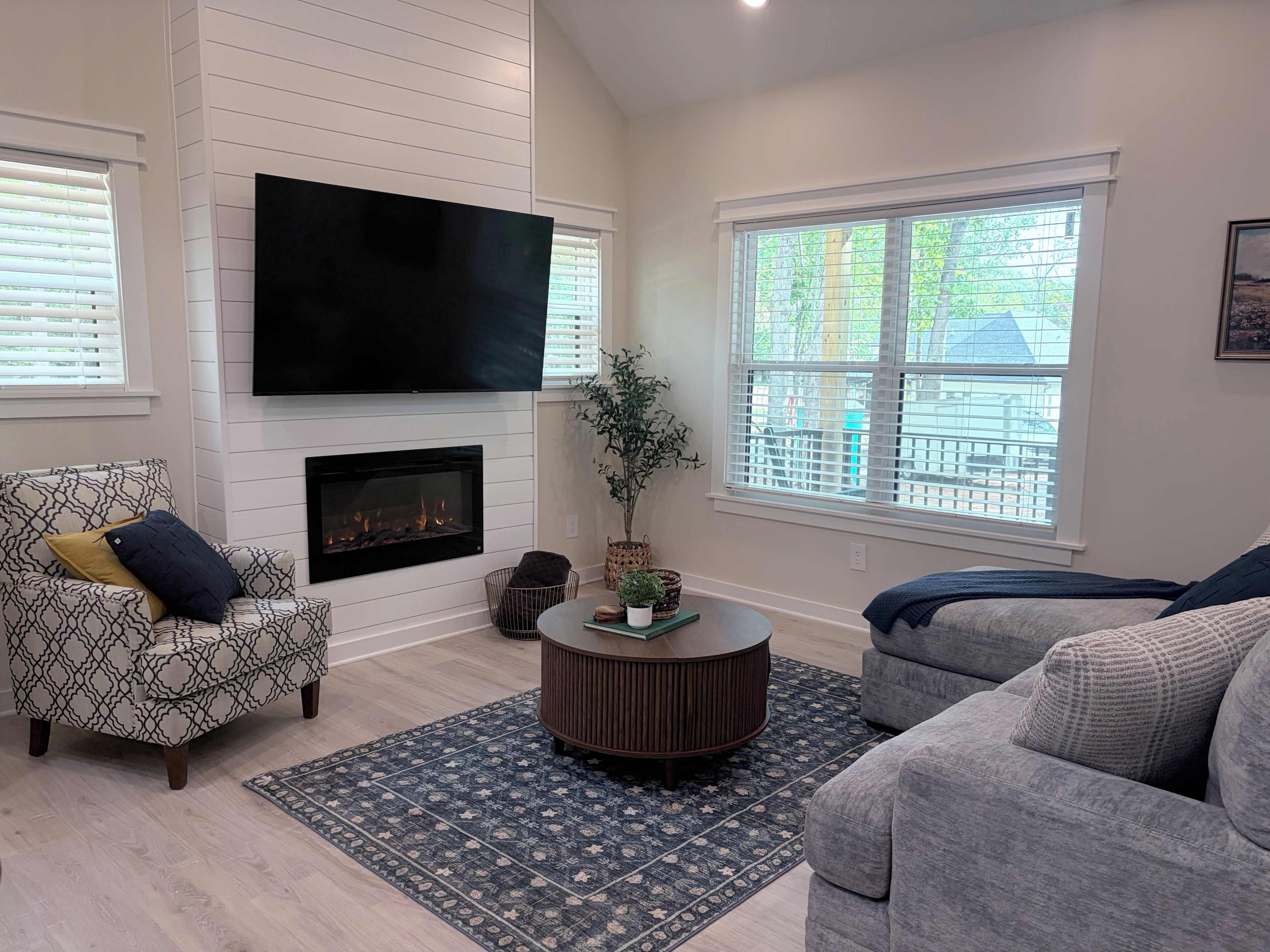 A living room with a fireplace, a flat-screen TV mounted above it, a patterned armchair, a round coffee table, and a large window with blinds.