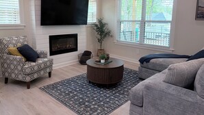 A living room with a fireplace, a flat-screen TV mounted above it, a patterned armchair, a round coffee table, and a large window with blinds.