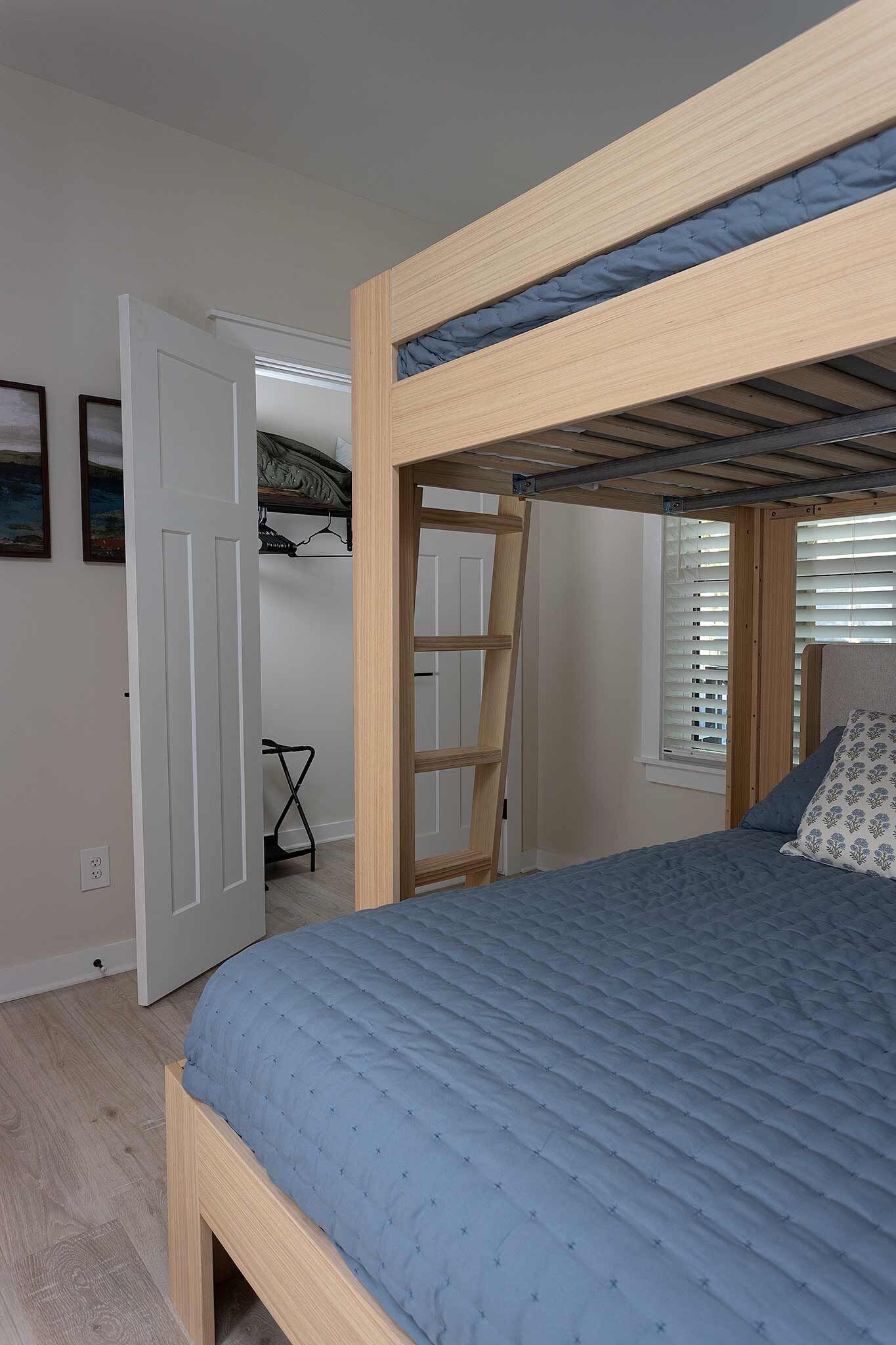 Un lit superposé avec une échelle, un matelas matelassé bleu, une tête de lit en bois et une fenêtre avec des stores.