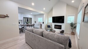 A modern living room with a grey sectional sofa, a flat-screen TV mounted above a fireplace, and a kitchen in the background.