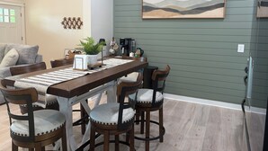 A dining area with a wooden table, chairs, a coffee machine, and a painting on the wall.
