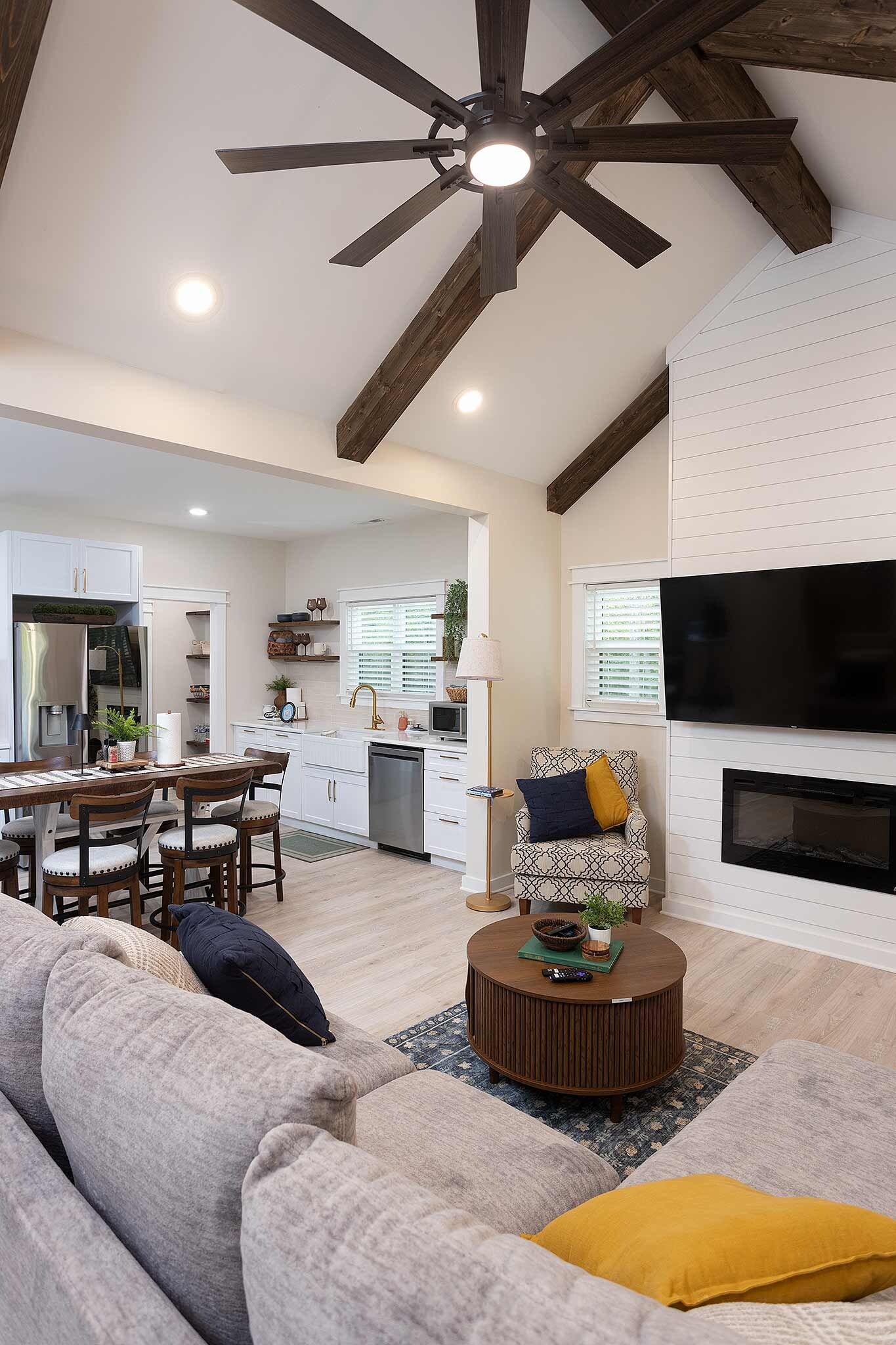 A modern living room with a ceiling fan, a flat-screen TV mounted on the wall, a dining area with a kitchen in the background, and a cozy seating area with a sofa and a coffee table.
