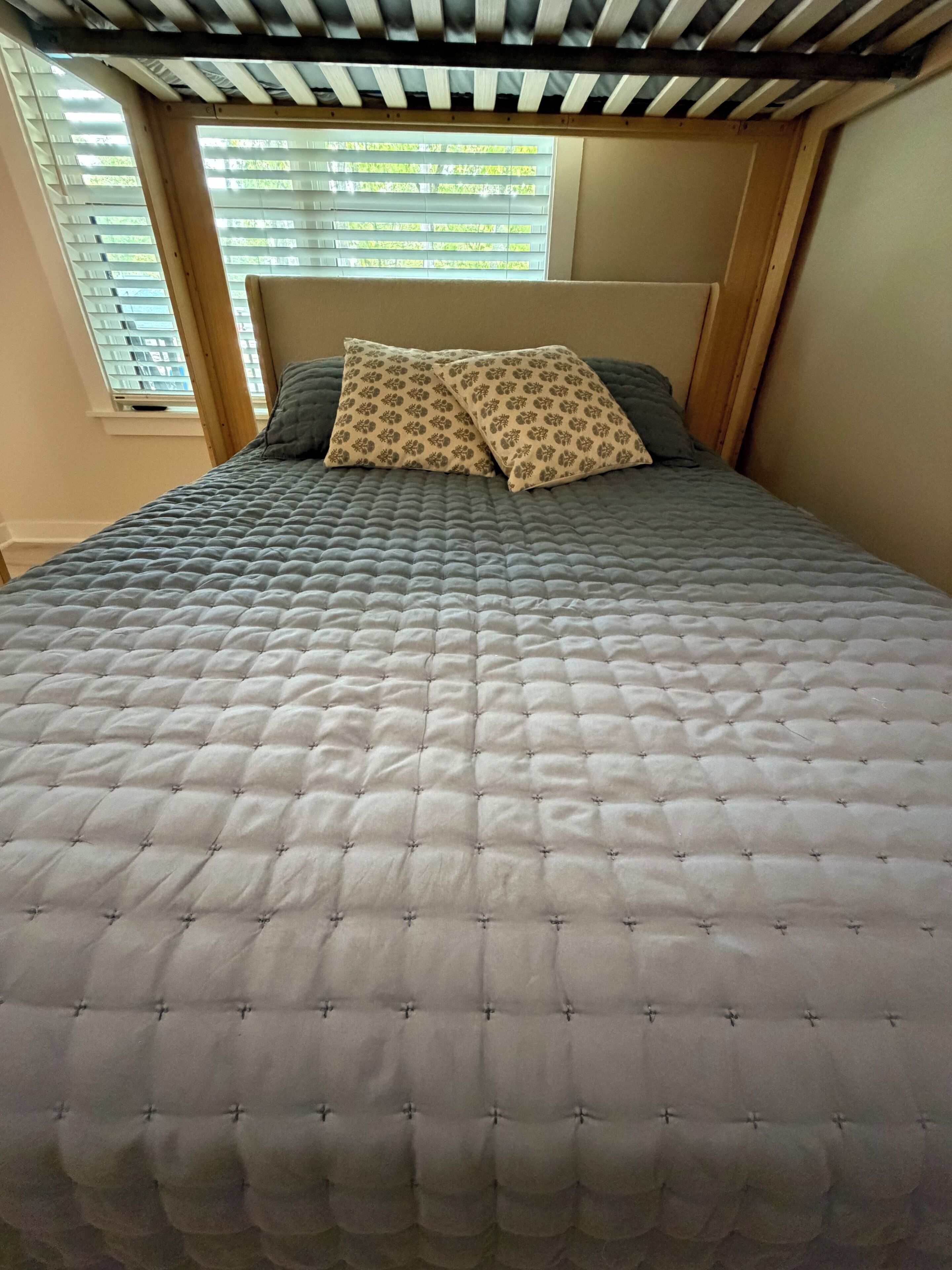 A bed with a quilted cover and two patterned pillows, a wooden headboard, and a window with blinds.