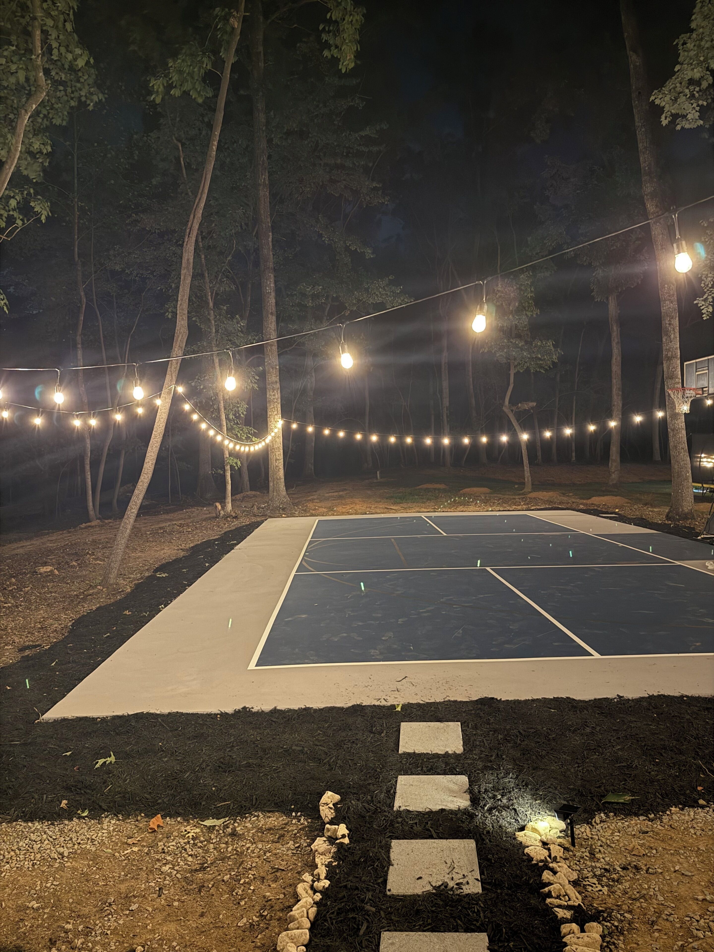 An outdoor basketball court with a net, surrounded by trees and illuminated by string lights.