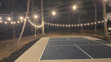 An outdoor basketball court with a net, surrounded by trees and illuminated by string lights.