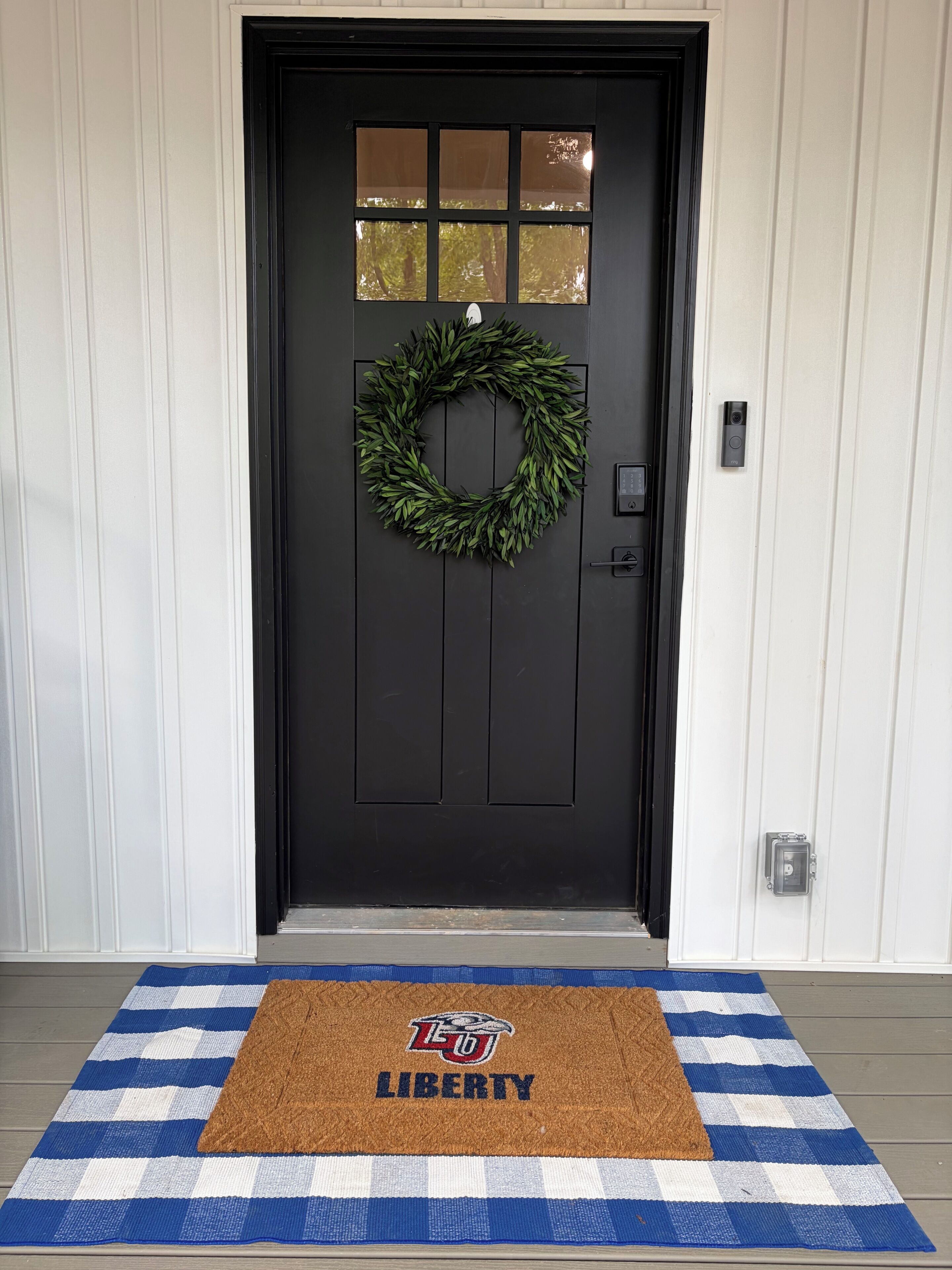 A black door with a wreath, a doorbell, and a blue and white checkered mat with 'LIBERTY' and a logo.