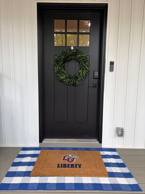 A black door with a wreath, a doorbell, and a blue and white checkered mat with 'LIBERTY' and a logo.