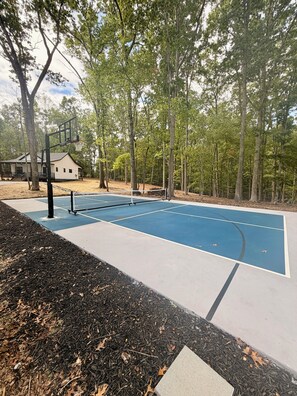 An outdoor basketball court with a net, surrounded by trees and a building in the background.