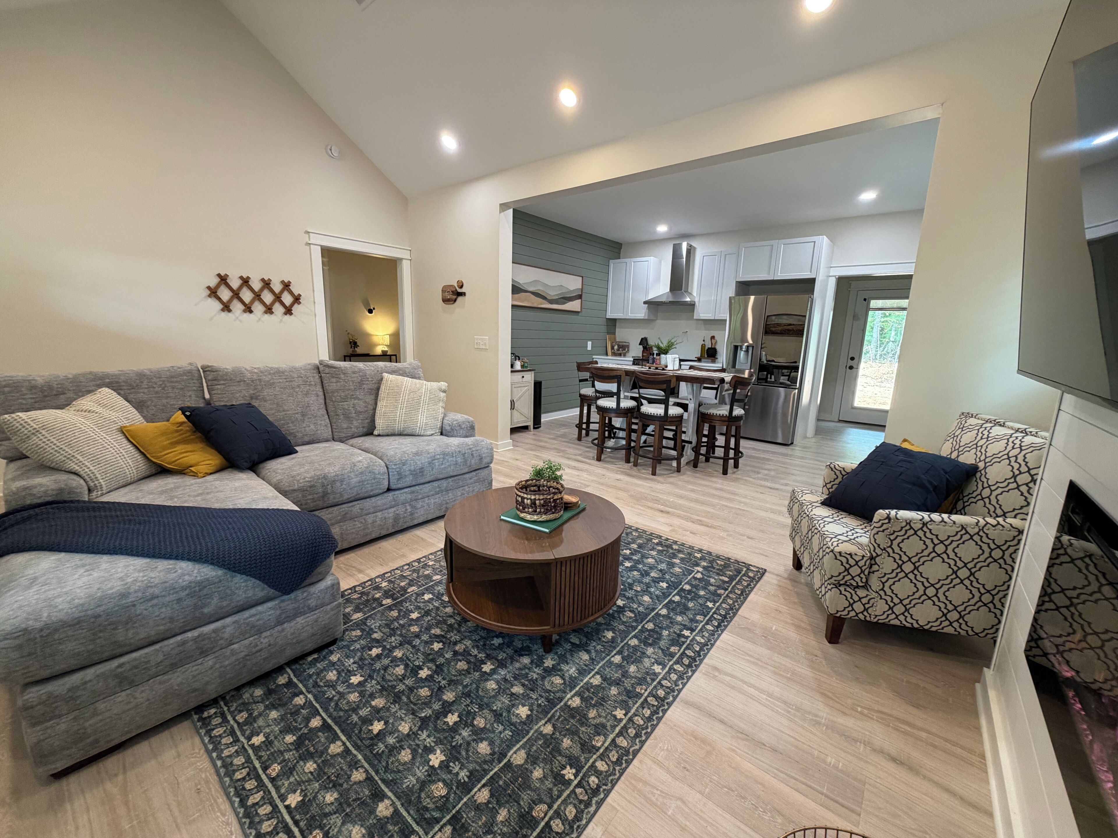 A modern living room with a grey sofa, patterned armchairs, a coffee table, and a rug. A kitchen with white cabinets and a stainless steel refrigerator is visible in the background.