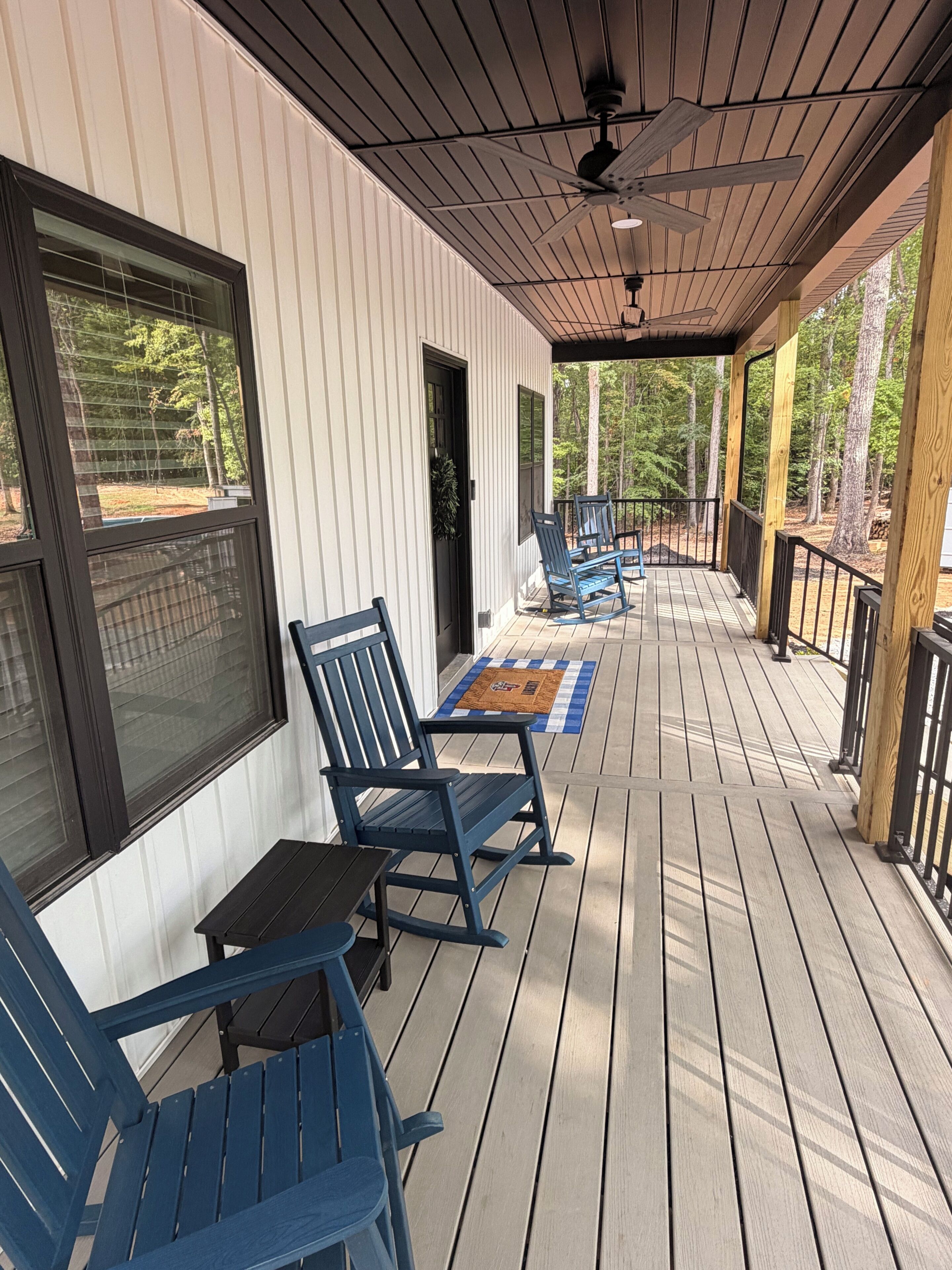 A covered porch with blue rocking chairs and a table, overlooking a wooded area.
