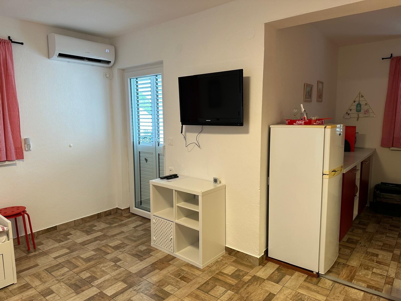 Une cuisine de taille réduite, avec un réfrigérateur blanc, un téléviseur fixé au mur et une fenêtre équipée de stores.