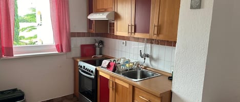 Cocina compacta con estufa a gas, horno y lavamanos. Hay una ventana con cortinas rojas, y el piso está cubierto de baldosas.