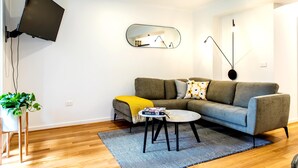Uma sala de estar moderna com um sofá cinzento, uma pequena mesa de centro, uma TV fixada na parede e um vaso com planta.