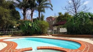 Uma piscina com borda de tijolos, cercada por vegetação e um edifício ao fundo.