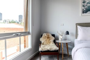 A hotel room with a large window, a plush chair, a small round table with a lamp, and a bed with white bedding. - Luxury spacious 2 Bedroom Fitzroy Apartment (Fitzroy)