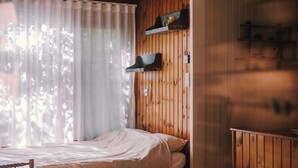 Quarto com cama, paredes de painel de madeira, janela com cortinas leves e uma estante com objetos.
