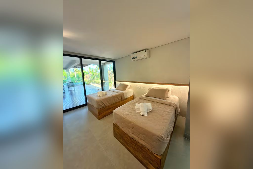 Une chambre d’hôtel moderne dotée de deux lits, d’une porte-fenêtre coulissante donnant sur l’extérieur et d’un appareil de climatisation fixé au plafond.