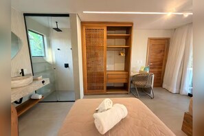 A hotel room with a bed, a wooden wardrobe, a bathroom with a glass shower enclosure, and a small seating area.