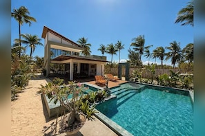 A modern house with a swimming pool, surrounded by palm trees and a sandy area.