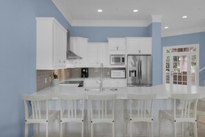 Cocina con armarios blancos, electrodomésticos de acero inoxidable y una barra con sillas blancas.