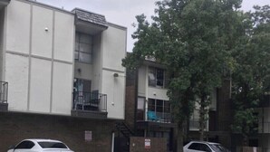 Un estacionamiento con dos autos, un edificio de fachada blanca y sección de ladrillo, y un árbol al fondo.