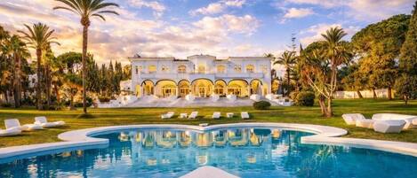 A pool with a white poolside area and a building with arches in the background.