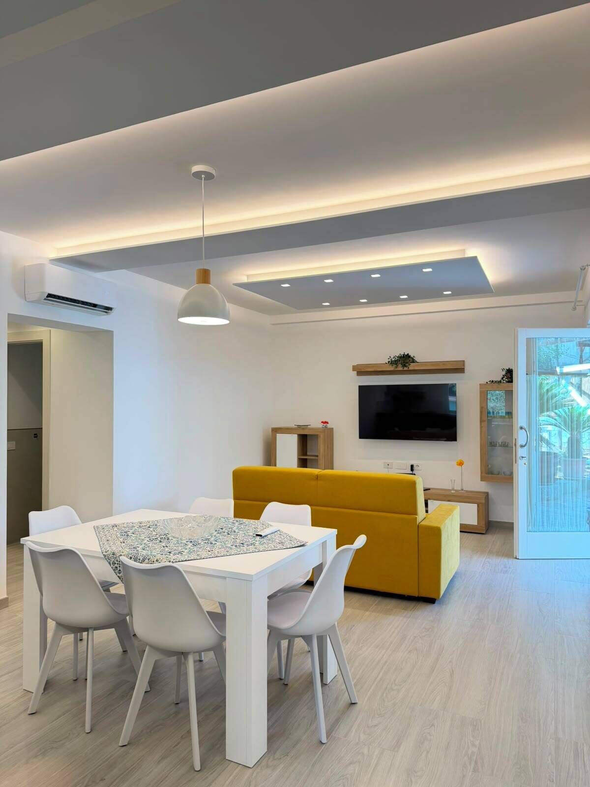 A modern living room with a dining area, a yellow sofa, a flat-screen TV, and a large window.