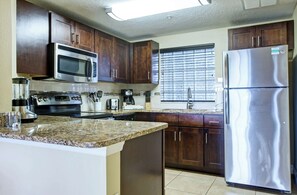 A kitchen with dark wooden cabinets, stainless steel appliances including a refrigerator, microwave, and stove, a granite countertop, and a window with blinds. - Cozy 2-bedroom condo with AC in lovely Clermont (Clermont)