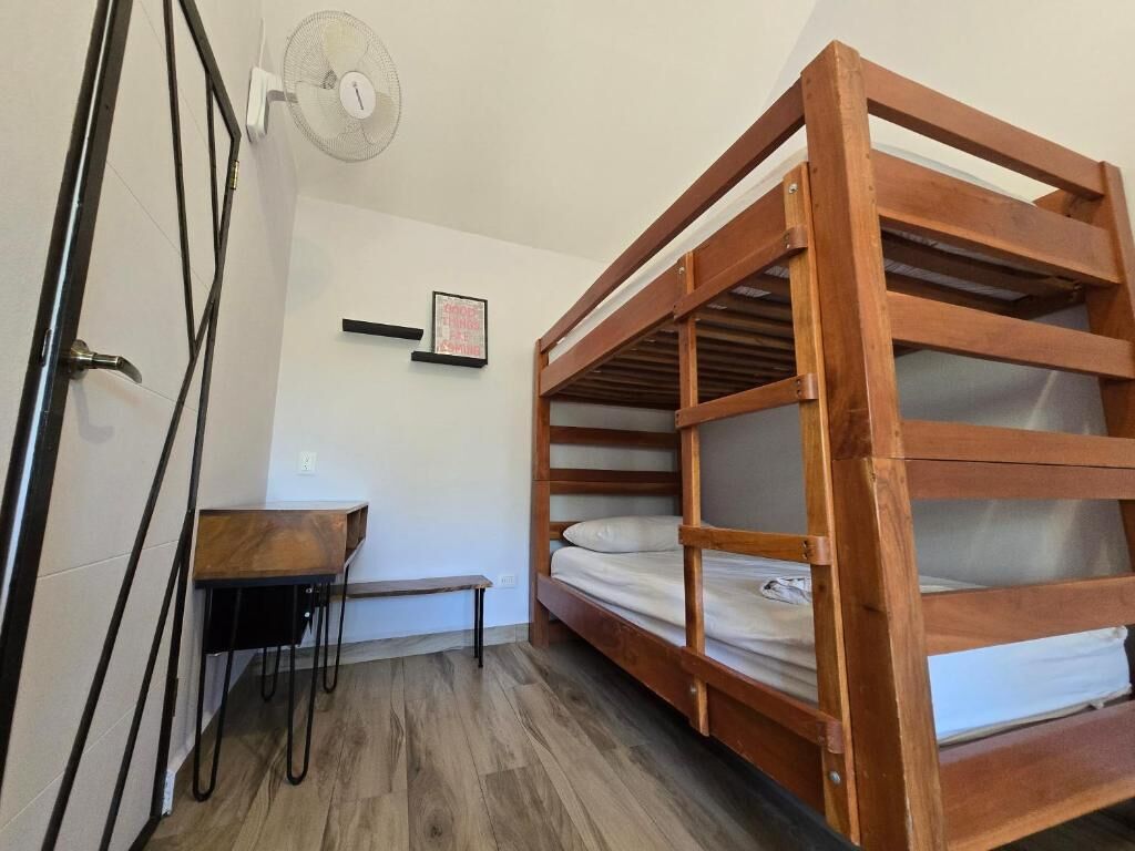 A bunk bed with a wooden frame and white bedding, a small table with a chair, a wall-mounted fan, and a framed picture.
