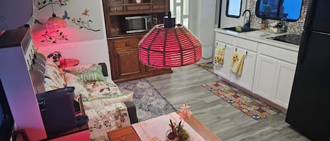 A compact living space with a kitchenette, a dining area with a table and chairs, a sofa, a red hanging lamp, and a television.