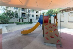 Ein Spielplatz mit Rutsche und Kletterwand, umgeben von einem überdachten Bereich und einem Gebäude im Hintergrund.