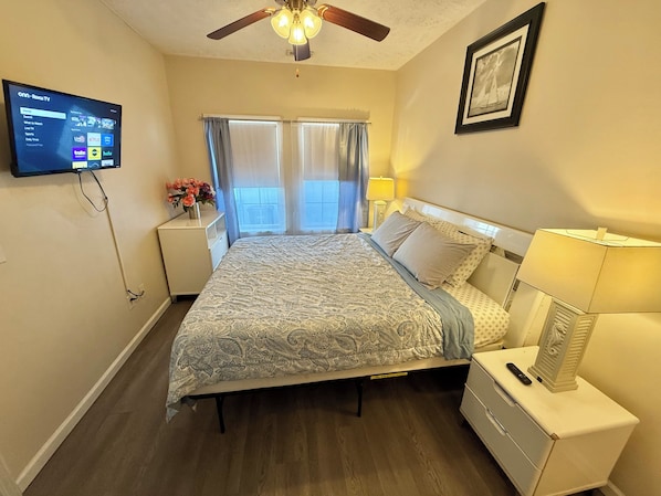Un dormitorio con cama, mesitas de noche, televisor montado en la pared, ventilador de techo y ventana con cortinas.