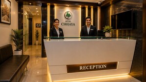 A hotel reception area with a reception desk, two staff members, a framed picture on the wall, and a potted plant. - Hotel Chhaya  (Chhatrapati Sambhajinagar)