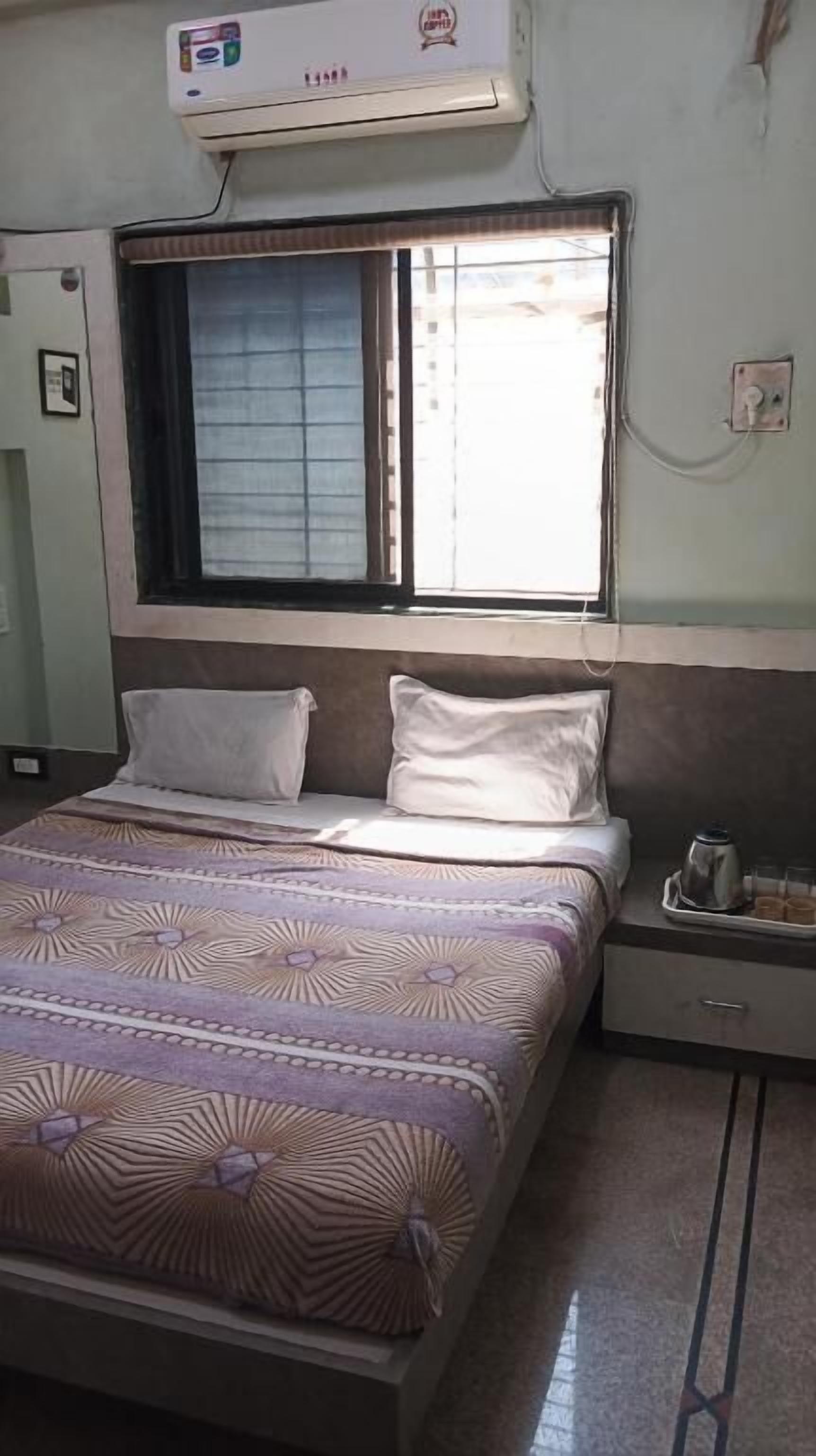 Habitación de hotel con cama, ventana con persianas, equipo de aire acondicionado y mesita de noche con una taza.