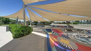 Un espacio al aire libre con sombra, un mural colorido, un sendero pavimentado y vegetación.