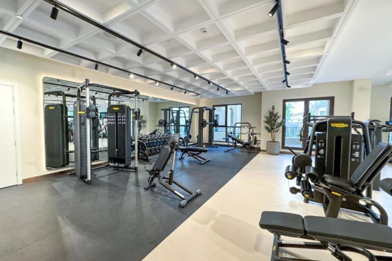 Et fitnesscenter med forskellig træningsudstyr, herunder styrkemaskiner og håndvægte, placeret i et åbent rum med store vinduer.