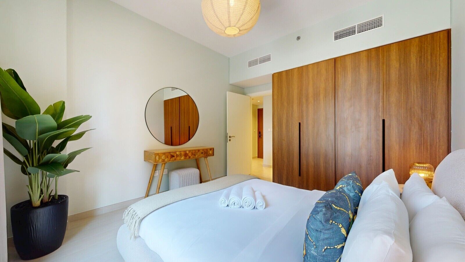A modern bedroom with a large bed, a wooden wardrobe, a round mirror, and a potted plant.