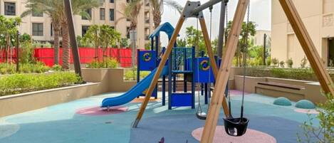 A playground with a slide and swings, surrounded by palm trees and a building in the background.