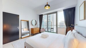 Ein Hotelzimmer mit einem großen Bett, einem Schminktisch, einem Spiegel, einem runden Spiegel, einem Fenster mit Vorhängen und Blick auf Gebäude.