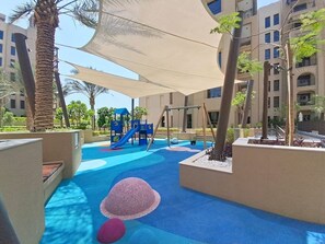 A children's play area with a slide and swings, surrounded by palm trees and shaded by a canopy. - Contemporary 1 Bed Near Burj Al Arab l Wild Wadi (Madinat Jumeirah Living)