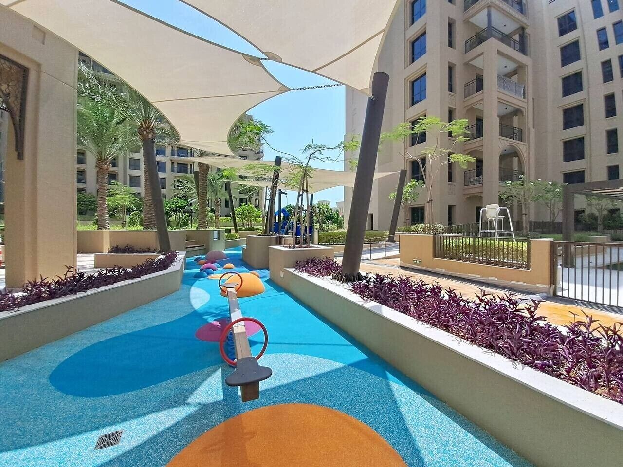 A children's play area with colorful ground surfaces, swings, and a shaded canopy.